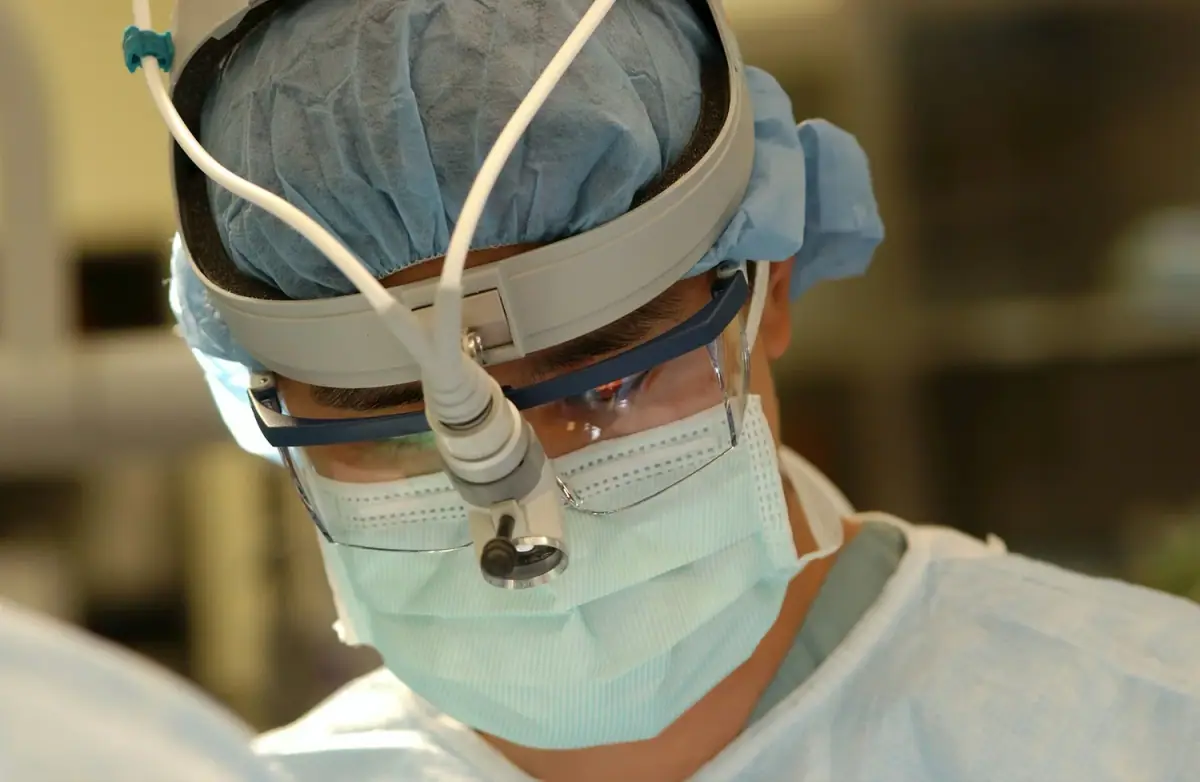 Close-up of a surgeon wearing a blue surgical cap, protective glasses, a face mask, and a headlamp, performing surgery.