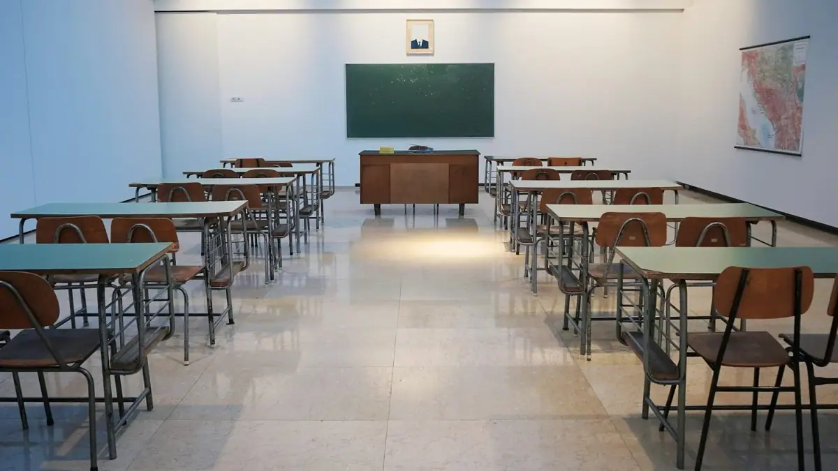 An empty classroom of desks and chairs facing a green chalkboard and a teacher’s desk.