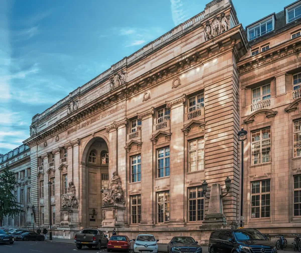 Historic neoclassical building with classical sculptures and ornate architectural details, with parked cars along the street in front.