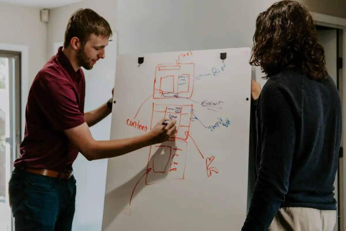 Two men discussing and outlining a website or app wireframe or diagram on a whiteboard, with notes and sketches.